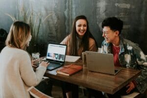 A photograph showing three people sat at a table, there are two open laptops on the table - the people at the table are smiling