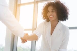 A woman shaking hands with a man, both are wearing white. There is a window behind the woman